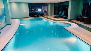 An indoor swimming pool with clear blue water, surrounded by lounge chairs and a cityscape view through large windows. - Bloom Hotel (Kuwait City)