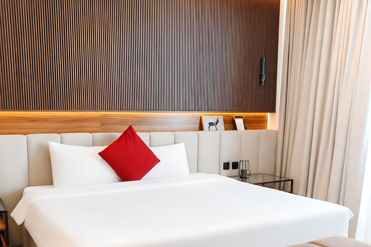 A modern hotel room with a large bed, a red accent pillow, a wooden headboard, and a bedside table with a lamp.