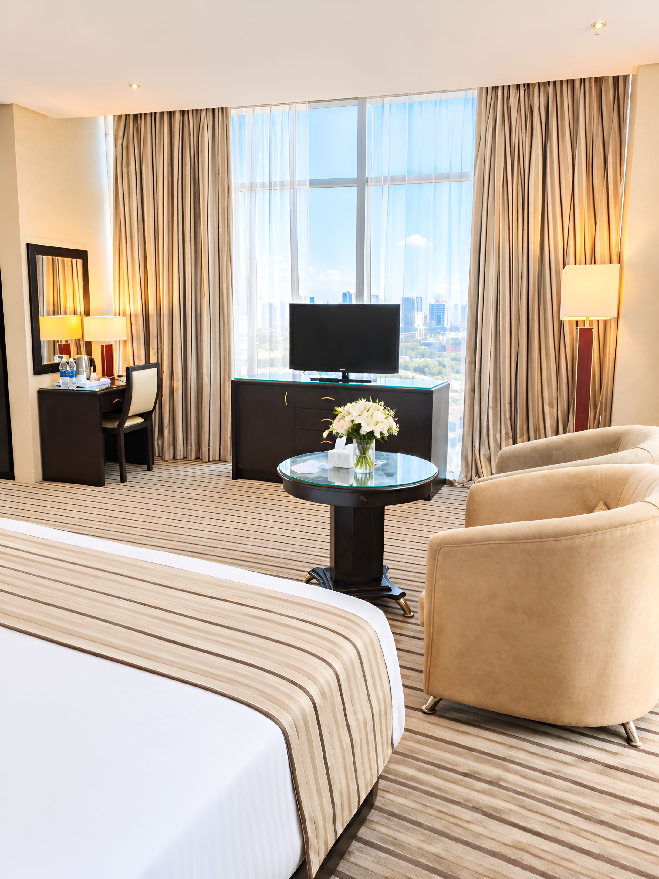 Habitación de hotel con una cama grande, un escritorio con silla, televisión y vistas de la ciudad a través de amplios ventanales.
