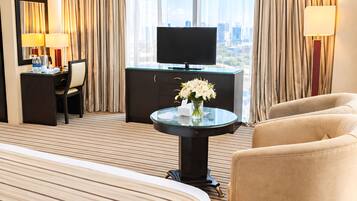 Habitación de hotel con una cama grande, un escritorio con silla, televisión y vistas de la ciudad a través de amplios ventanales.