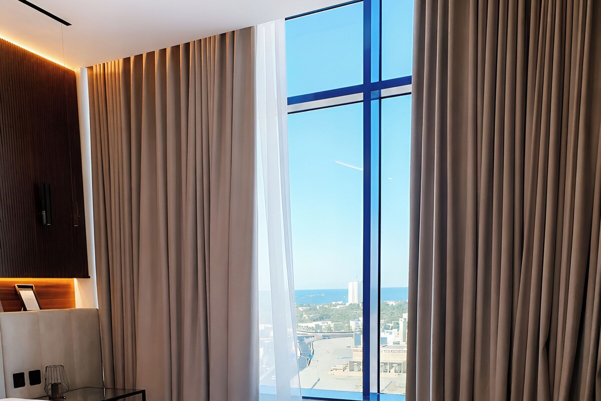A modern hotel room with a large bed, a seating area, and a view of the cityscape through large windows.