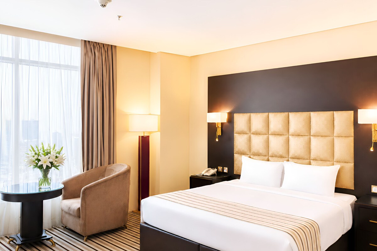 Habitación de hotel con una cama grande, una silla, una mesita de noche, una lámpara y un ventanal con cortinas.
