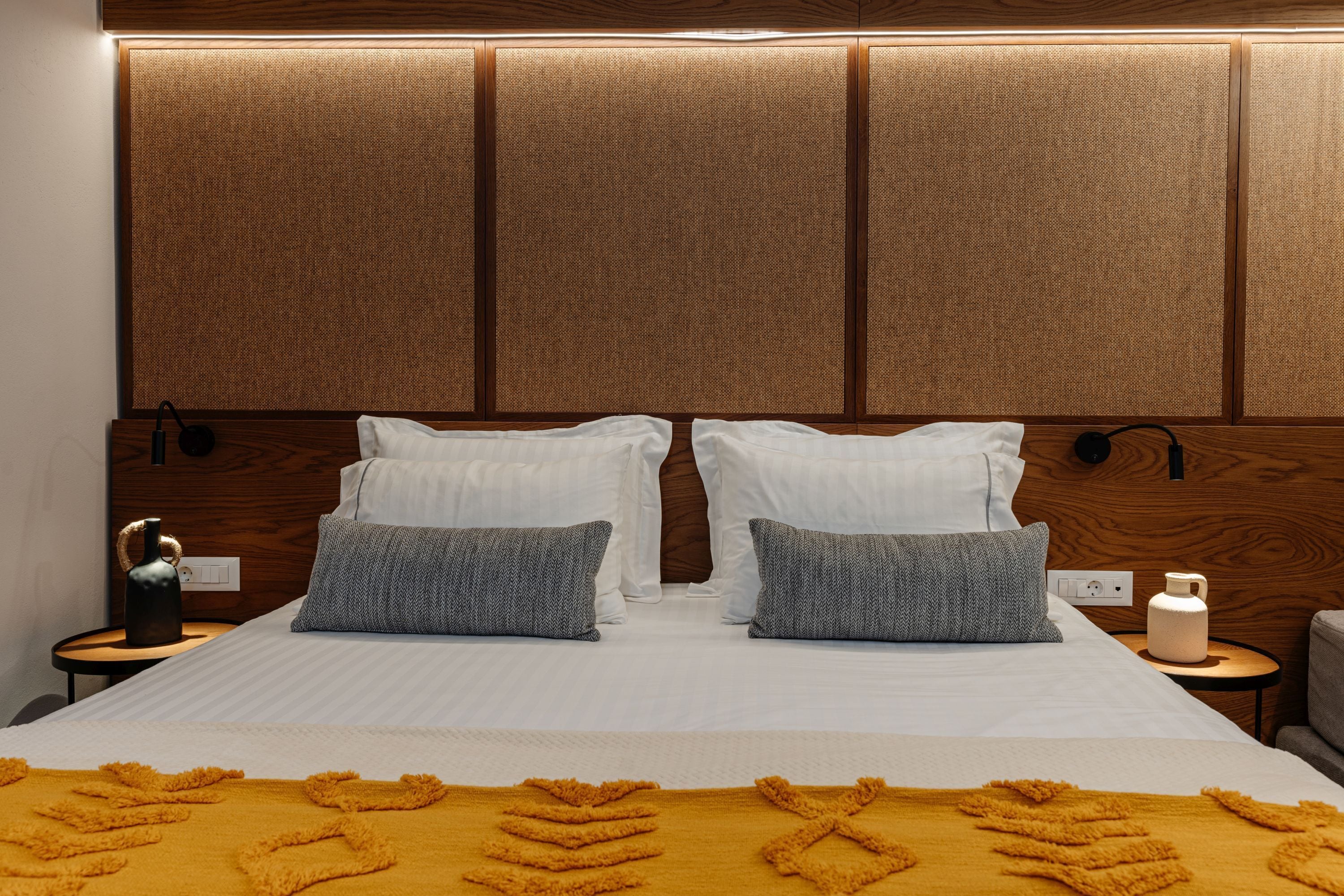 Habitación de hotel con una cama grande, dos almohadas, una mesita de noche con lámpara y un panel en la pared sobre la cama.