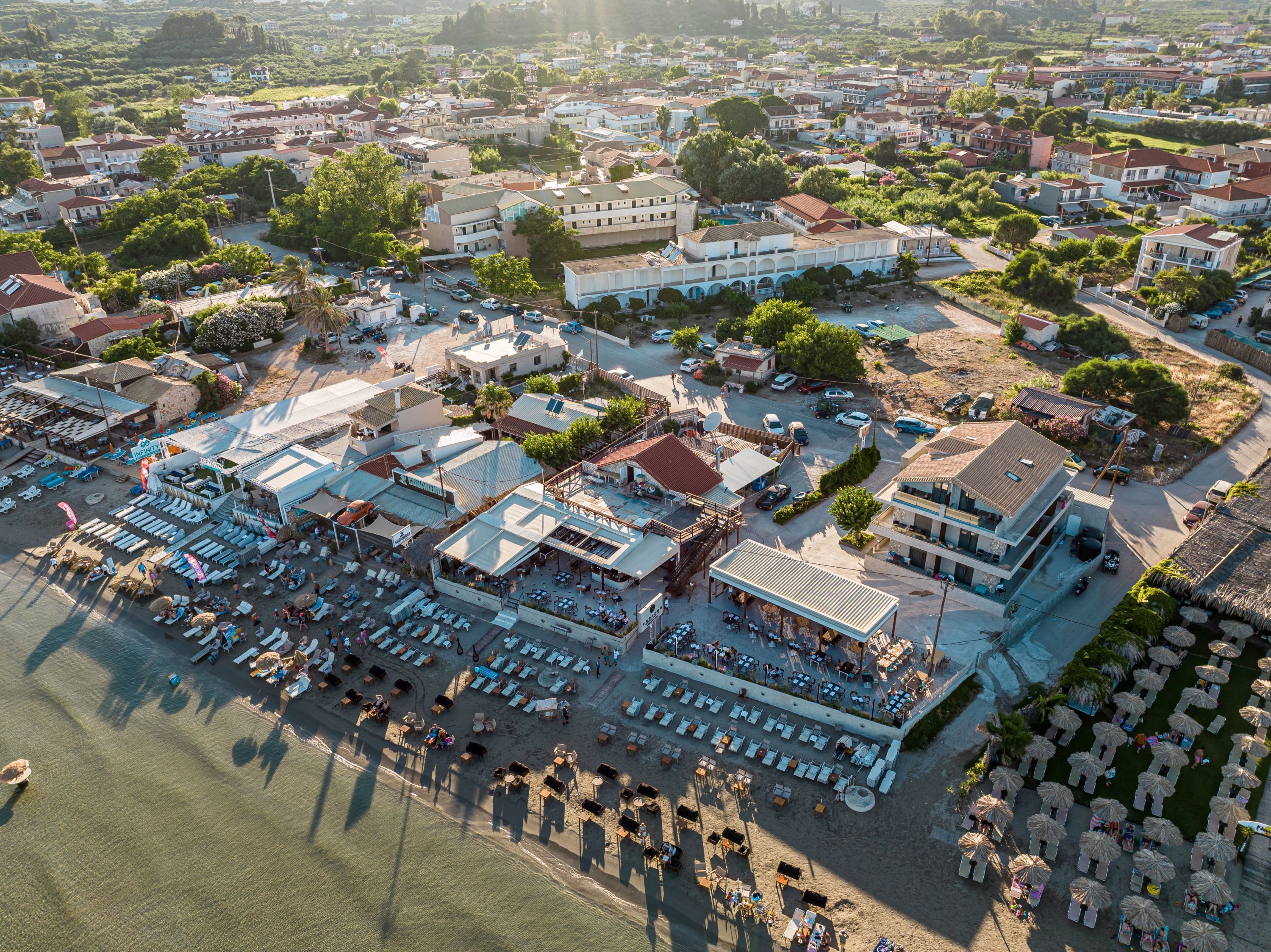 An aerial view of a coastal town with a beachfront area featuring multiple buildings, including a prominent white building with a blue roof, and several beachside cafes and restaurants.