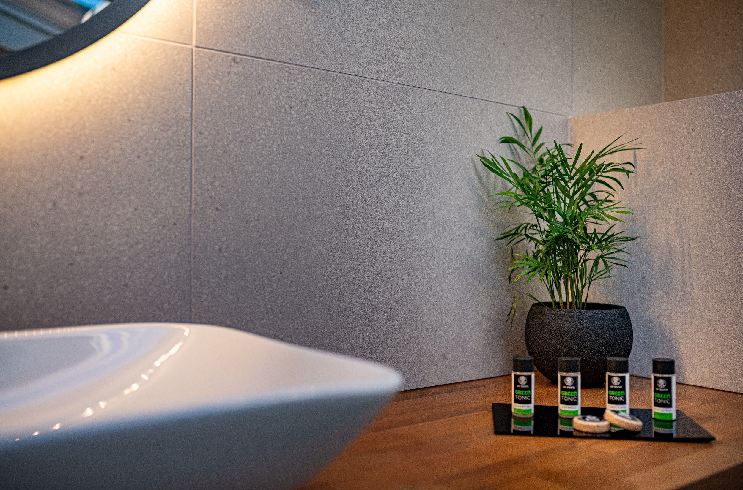 A bathroom with a white sink, a potted plant, and three bottles of green tonic on a wooden tray.