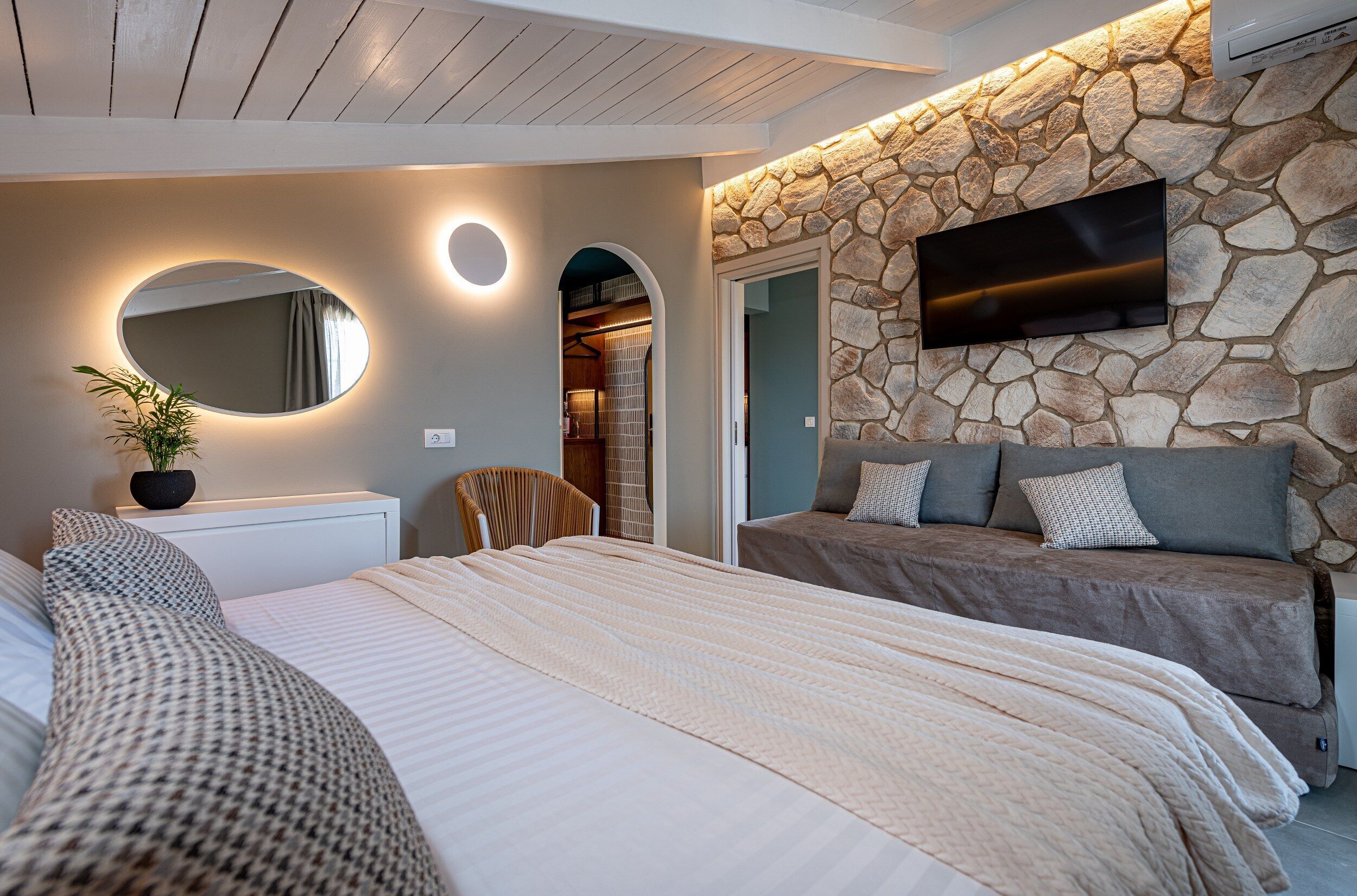 Un dormitorio con una cama grande, un espejo redondo, una pared de piedra y un televisor montado en la pared.