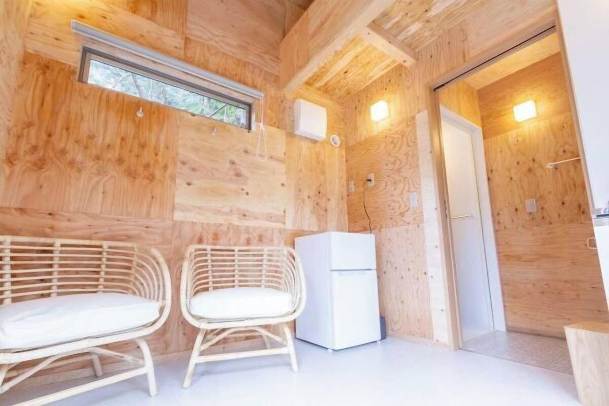 L’intérieur d’un chalet en bois, avec deux fauteuils en osier, un réfrigérateur blanc et une porte donnant sur une autre pièce.