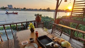 A wooden table set for dining with a river view, a grill, and a boat in the background.