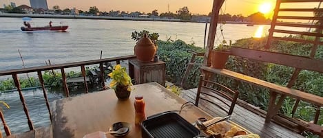 A wooden table set for dining with a river view, a grill, and a boat in the background.