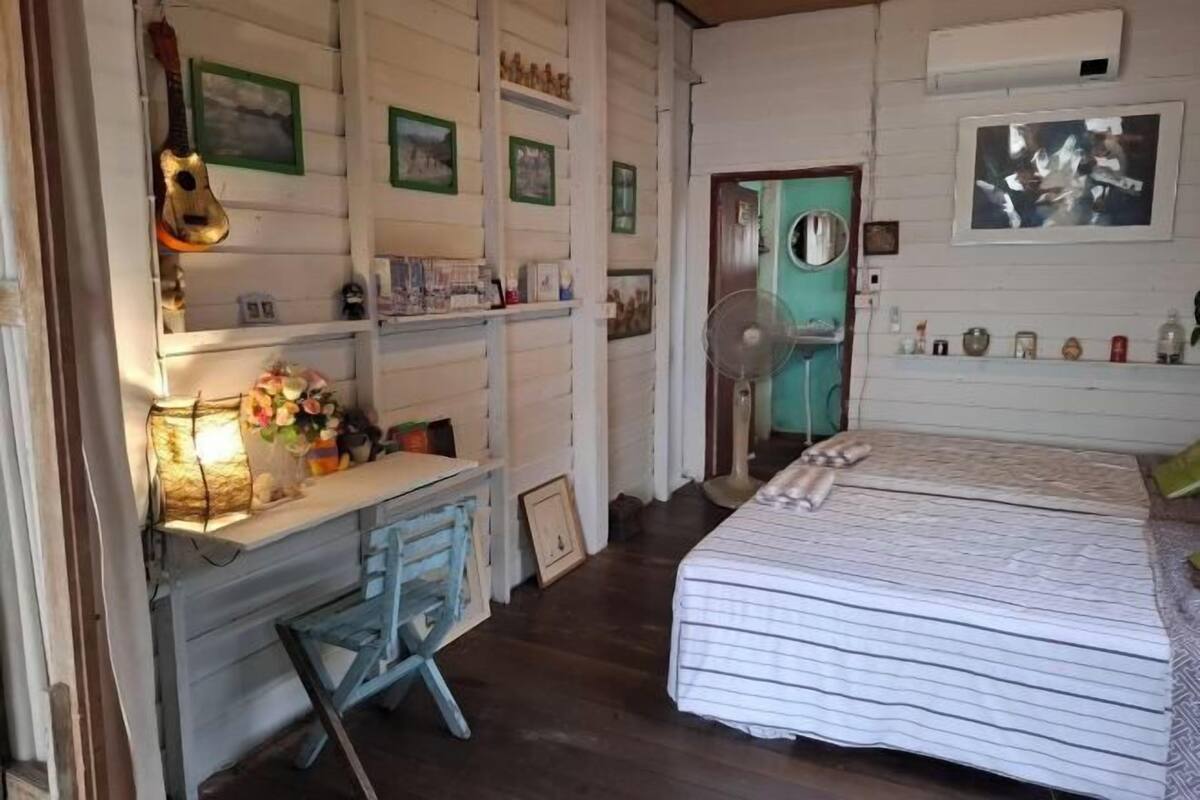 A room with a bed, a desk with a chair, a fan, and wall-mounted shelves with various items.