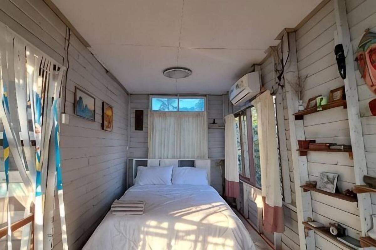 A narrow room with a bed, a window with curtains, a ladder, and shelves with various items.