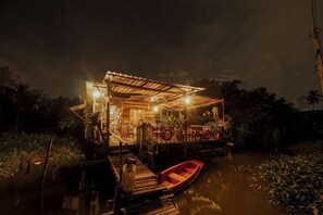 A houseboat with a covered deck, a small boat docked in front, and a water body with vegetation around. - Baan GoLite Ko Kret (Pak Kret)