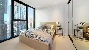 Ein modernes Schlafzimmer mit großem Fenster, einem Bett mit gemusterten Betttextilien, Nachttischen mit Lampen und Blick auf hohe Gebäude.
