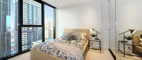 Une chambre moderne avec une grande fenêtre, un lit recouvert d’une literie à motifs, des tables de chevet avec des lampes, et une vue sur des immeubles de grande hauteur.