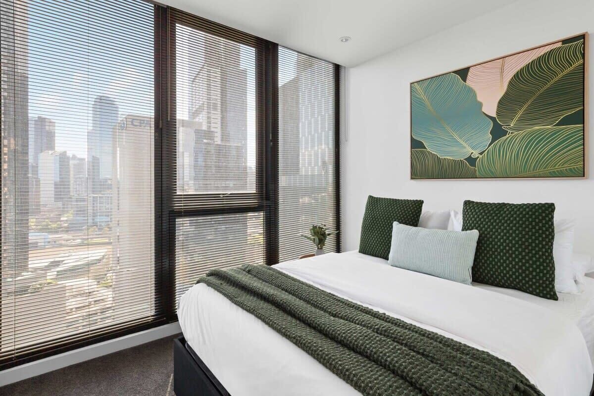 A modern bedroom with a large bed, city view through floor-to-ceiling windows, and a green abstract painting on the wall.