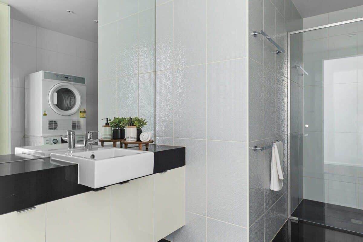 A modern bathroom with a built-in washing machine, a large mirror, and a glass shower enclosure.