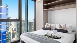 Habitación de hotel moderna con un ventanal grande que ofrece vistas a la ciudad, una cama bien hecha con almohadas y una bandeja con una toalla y plantas.