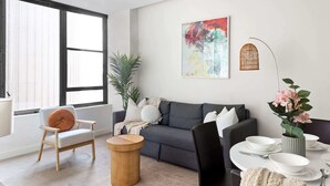 Een moderne woonkamer met een grijze bank, een houten stoel, een rond bijzettafeltje, en een eethoek met witte borden en kommen.