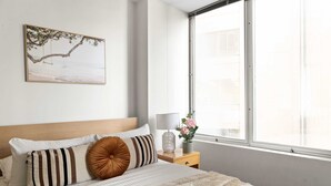 Een keurig opgemaakt slaapkamer met een bed, kussens, een nachtkastje met een lamp, een vaas met bloemen en een raam met uitzicht op zee.
