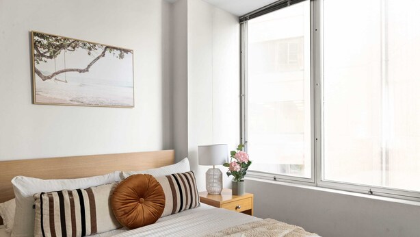 Een keurig opgemaakt slaapkamer met een bed, kussens, een nachtkastje met een lamp, een vaas met bloemen en een raam met uitzicht op zee.