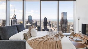 Quarto de hotel moderno com uma cama grande, um sofá e vista para a cidade através de janelas do chão ao teto.