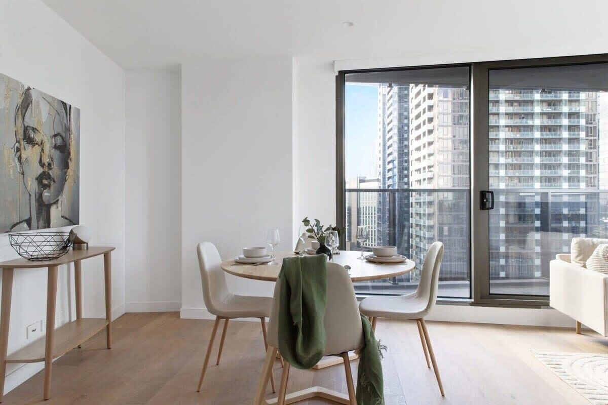 A modern dining area with a round table, white chairs, and a view of tall buildings through large windows.