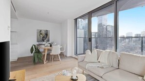 A modern living room with a sofa, a dining table, and chairs. There is a large window offering a view of the cityscape.