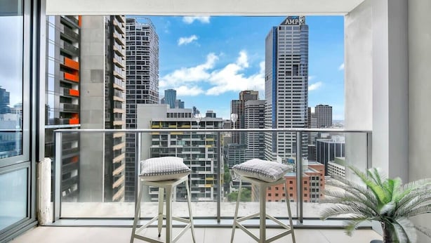 A balcony with two white stools and a potted plant, overlooking a cityscape with tall buildings. - Comfy 2BR Apt Near Southern Cross With City Views (Melbourne)