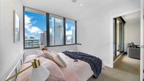 A modern bedroom with a large window, a bed with a striped bedspread, a bedside lamp, and a view of the city skyline. - Comfy 2BR Apt Near Southern Cross With City Views (Melbourne)