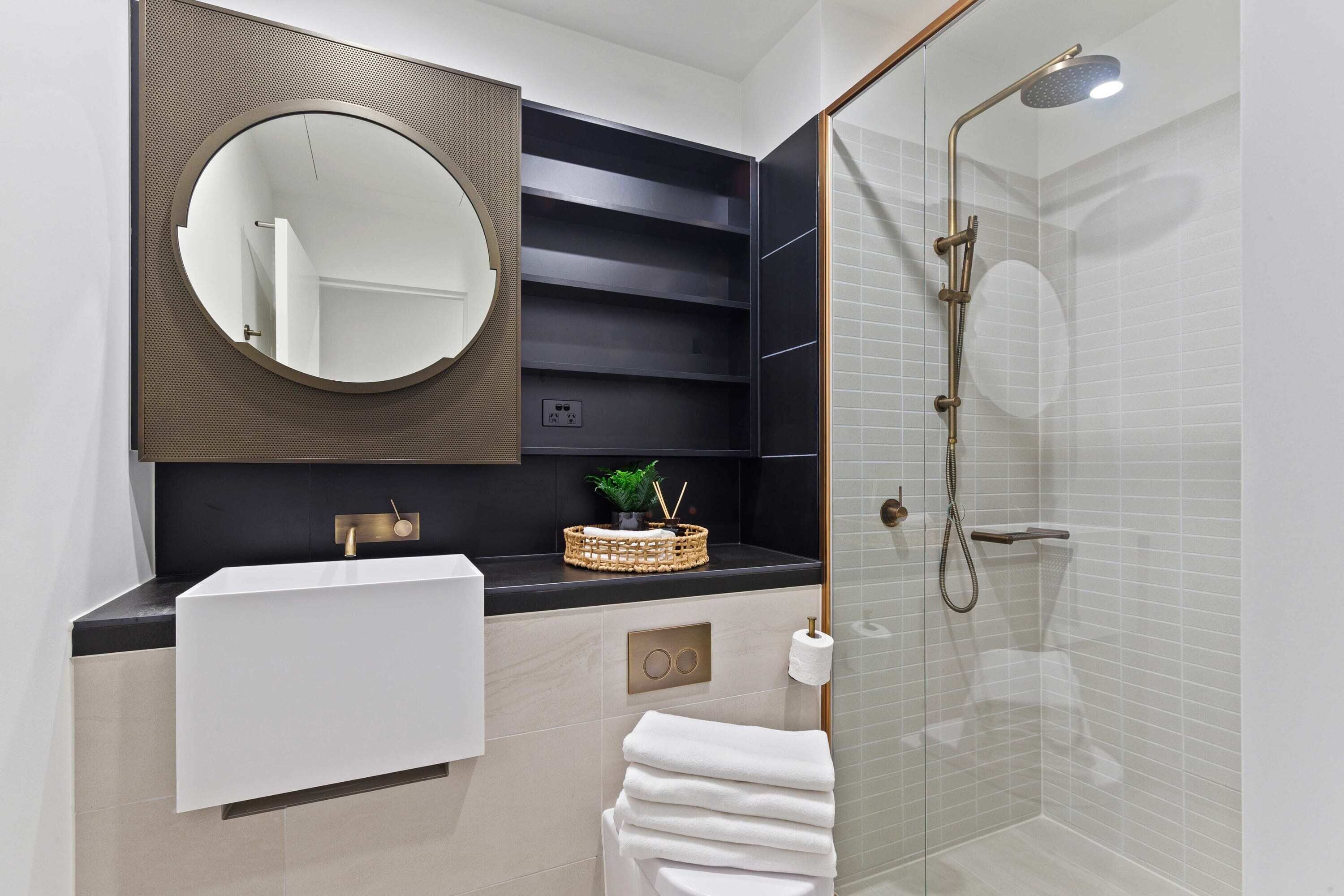 A modern bathroom with a round mirror, a white sink, a glass-enclosed shower, and a black shelving unit.