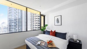 A modern hotel room with a large window, a bed with a gray and white striped blanket, a small bedside table with a lamp, and a framed picture on the wall. - A Chic 2BR Apt, City Views Near Southern Cross (Melbourne)