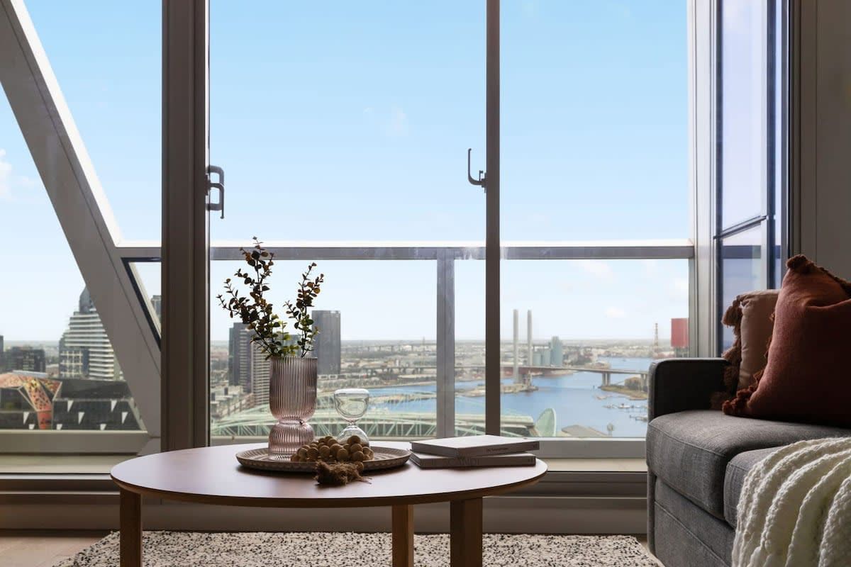 A modern living room with a large window offering a city view, a round wooden table with a vase and decorative items, and a grey sofa with cushions.