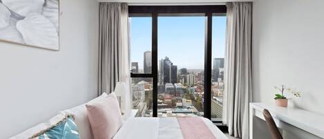 Ett modernt hotellrum med en stor säng, ett skrivbord och utsikt över stadsbilden genom stora fönster från golv till tak.
