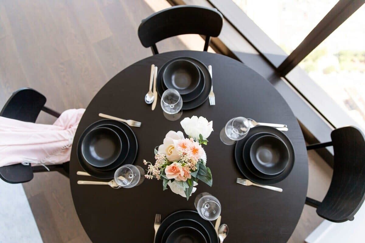 A round table set for four with black plates, silverware, and a floral centerpiece.