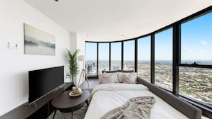 A modern hotel room with a curved glass wall offering a city view, a flat-screen TV, a bedside table with a lamp, and a large bed with a blanket. - Lovely 3BR Apt With Panoramic Views, Free Parking (Melbourne)