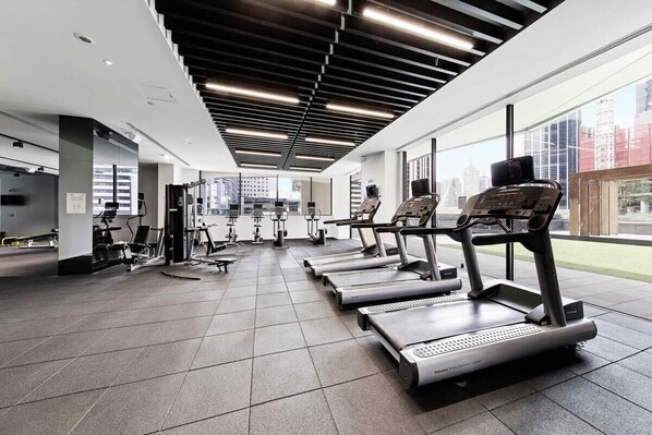 Une salle de sport moderne équipée de tapis de course, d’appareils de musculation et dotée d’une grande fenêtre offrant une vue sur la ville.