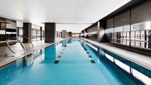 A modern indoor swimming pool with clear glass walls, a long poolside area, and a view of the cityscape.