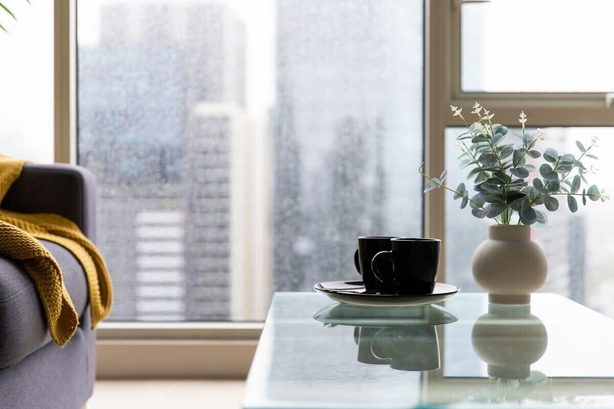 A room with a view of a cityscape through a window, a black cup on a glass table, and a vase with green foliage.