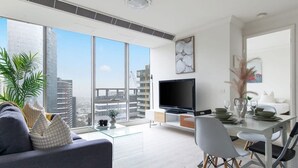 A modern living room with a large TV, dining area, and large windows with a city view.