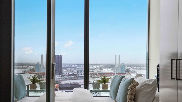 A modern living room with a large window offering a city view, a sofa with cushions, and two potted plants. - Southern Cross Gem: Modern Home With harbour Views (Melbourne)