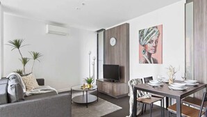 Una sala de estar moderna con un sofá gris, una mesa redonda de centro, un televisor de pantalla plana y un comedor con una mesa y sillas de madera.