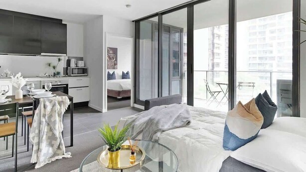 Habitación de hotel moderna con una cama grande, una mesa de comedor de vidrio, una zona de cocina y un balcón con vistas a la ciudad.
