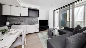 A modern kitchen with white cabinets, a marble backsplash, and a black countertop. There is a dining table with chairs and a small side table. A large window provides a view of the city.