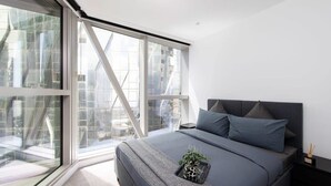 A modern bedroom with a large window, a bed with grey bedding, and a small plant on the bed.