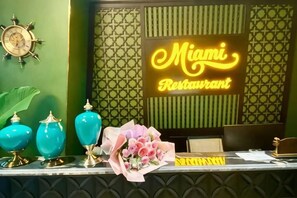 Un comptoir d'accueil avec un panneau néon indiquant « Miami Restaurant », une horloge et des vases décoratifs.