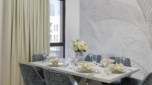 A dining table set for six with white plates, silverware, and clear glasses, flanked by grey upholstered chairs. A vase with white flowers is on the table, and a large abstract wall art is behind it.