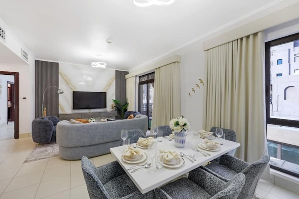 A modern living room with a dining area set for a meal, a grey sofa, and a flat-screen TV mounted on the wall.