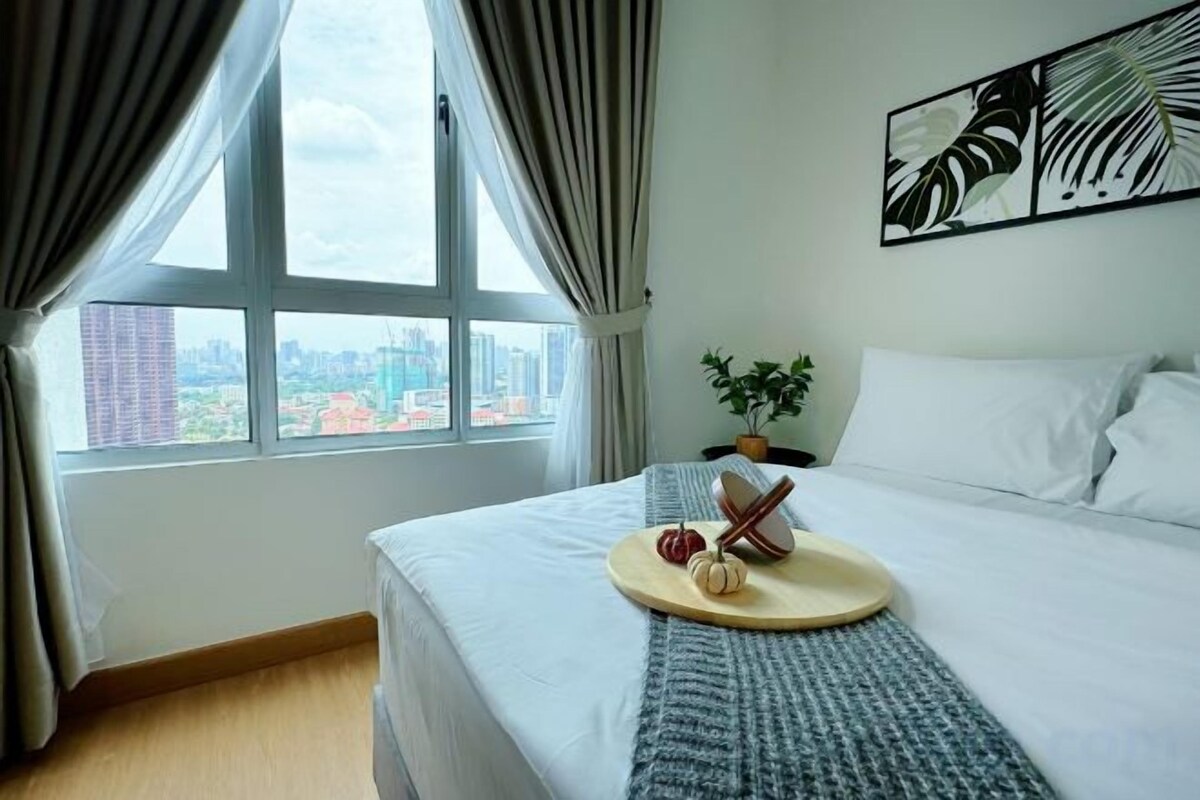 A hotel room with a bed, a window with curtains, a city view, a plant, and a painting on the wall.