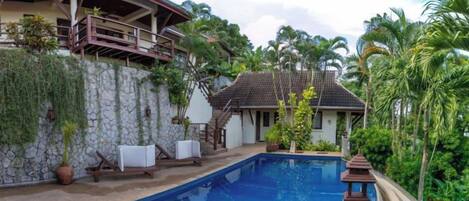 Una piscina con un muro de piedra y una cubierta de madera, rodeada de abundante vegetación y una casa con balcón.