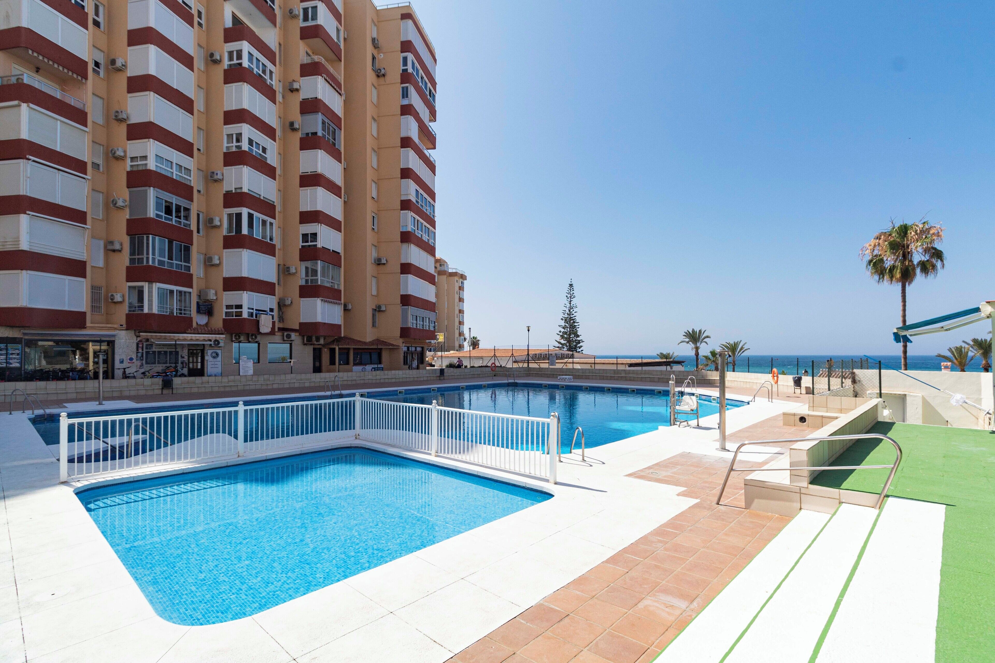 A swimming pool with a white fence, a palm tree, and a building with balconies in the background.